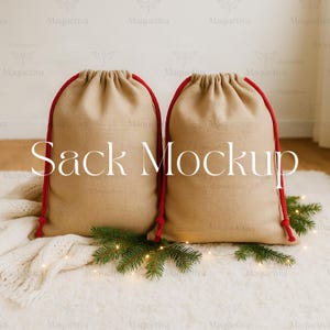 May include: Two beige burlap sacks with red drawstrings, arranged side-by-side. The text "Sack Mockup" is displayed in white. The sacks rest on a white, textured surface, adorned with pine branches and small lights.