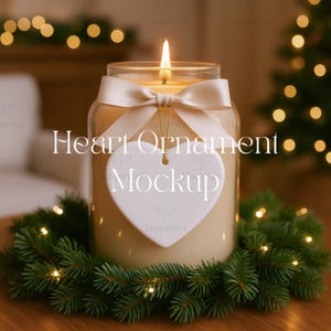 May include: A lit candle in a glass jar with a cream-colored ribbon and a heart-shaped ornament. The candle sits on a bed of green pine needles with small lights. The text "Heart Ornament Mockup" is on the image.