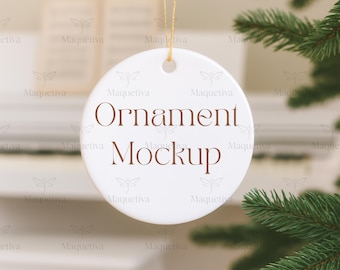May include: A white, round ornament with the words "Ornament Mockup" in brown script. The ornament hangs from a gold string, with a blurred background and a sprig of green pine needles in the corner.