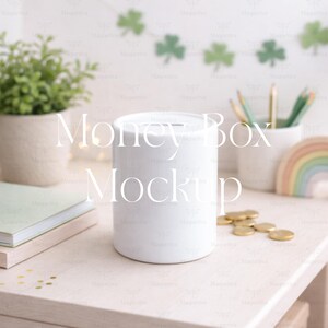 May include: A white cylindrical money box with a slot on top, sitting on a light pink surface. Gold coins are scattered nearby. A small potted plant, a notebook, and a rainbow decoration are also visible.
