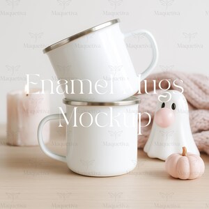 May include: Two white enamel mugs with silver rims are stacked. A pink candle, a ghost-shaped figurine with a bubblegum nose, and a small pink pumpkin are arranged nearby. The text "Enamel Mugs Mockup" is overlaid on the image.