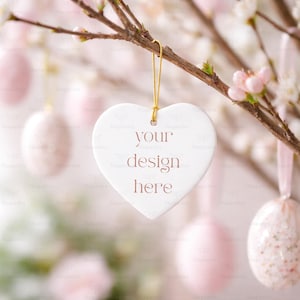 Puede incluir: Un adorno blanco en forma de corazón con el texto "your design here" cuelga de un hilo dorado. El adorno está suspendido de una rama de árbol con flores rosas y huevos de Pascua en el fondo.