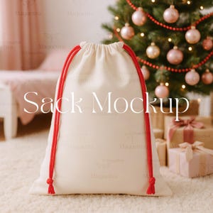 May include: A cream-colored drawstring sack with red rope ties. The text "Sack Mockup" is displayed on the front of the bag. The background includes a Christmas tree with pink and gold ornaments and wrapped gifts.