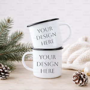 May include: Two white enamel mugs stacked vertically, each featuring a black rim and the black text "YOUR DESIGN HERE" printed on the front. The mugs are positioned on a light wooden surface with pine branches and a white knit fabric nearby.