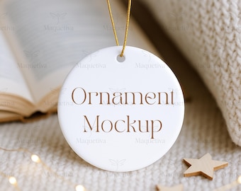 May include: A white, round ornament with the words "Ornament Mockup" in brown script. The ornament hangs from a gold string. The background includes a book, a knitted blanket, and wooden star decorations.