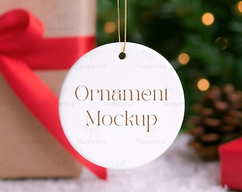 Könnte beinhalten: Ein weißer, runder Ornament mit dem Schriftzug "Ornament Mockup" in braunen Buchstaben, der an einer goldenen Schnur hängt. Im Hintergrund ein Geschenk mit rotem Band, sowie ein Weihnachtsbaum und ein Tannenzapfen.