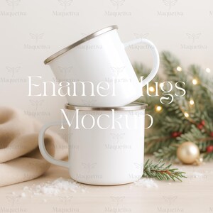 Könnte beinhalten: Zwei weiße Emaille-Tassen mit silbernen Rändern sind auf einer hellen Holzoberfläche gestapelt. Die Tassen haben gebogene Griffe und der Text "Enamel Mugs Mockup" ist auf dem Bild überlagert. Ein beiger Schal, Tannenzweige und eine goldene Kugel befinden sich im Hintergrund.