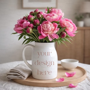 May include: A white ceramic jug with a handle, filled with bright pink peonies and green foliage. The jug has the text "your design here" in a soft pink colour. The arrangement sits on a wooden tray with a small white bowl and a folded beige napkin.