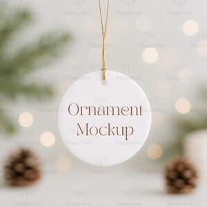 May include: A white, round ornament mockup with the text "Ornament Mockup" in brown. The ornament hangs from a gold string. The background features blurred lights, pine needles, and pine cones.