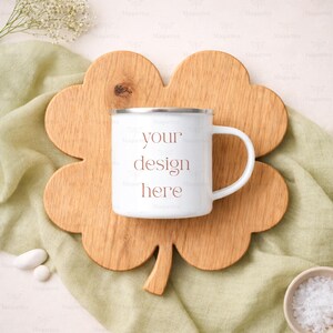 May include: A white enamel mug with a silver rim and a white handle, featuring the text "your design here". The mug sits on a wooden, clover-shaped cutting board. The background includes a green cloth and small white decorative elements.