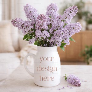 May include: A white ceramic jug with a handle, displaying the text "your design here" in a soft brown hue. The jug contains a bouquet of purple lilacs. Some lilac sprigs are on the table.