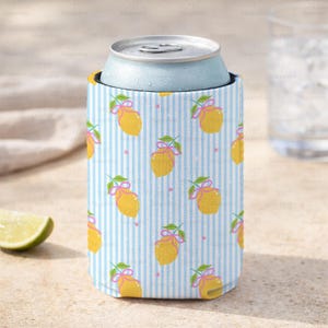 May include: A blue and white striped can cooler featuring a pattern of yellow lemons with pink bows and green leaves. A light blue can sits inside the cooler. A lime wedge is in the lower left corner.