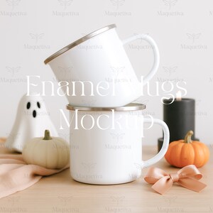 May include: Two white enamel mugs stacked on a wooden surface, with a ghost figurine, a small pumpkin, and a black candle. A peach-colored bow and a second pumpkin are also present. The mugs have silver rims.