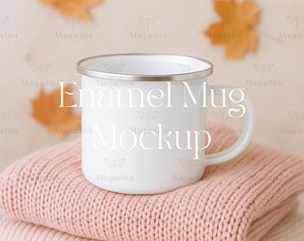 May include: A white enamel mug with a silver rim sits atop a stack of two pink knitted sweaters. The mug has the words "Enamel Mug Mockup" printed on it. Autumn leaves are in the background.