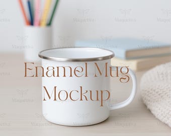 May include: A white enamel mug with a silver rim and handle, featuring the text "Enamel Mug Mockup" in brown. The mug is on a light-colored wooden surface, with pencils and books in the background.