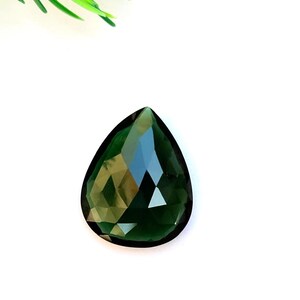 Op de afbeelding: Een gefacetteerde, druppelvormige edelsteen in een diepe bosgroene kleur. De edelsteen heeft een gepolijst oppervlak met meerdere facetten die licht reflecteren. Groen gebladerte is zichtbaar in de linkerbovenhoek, tegen een witte achtergrond.