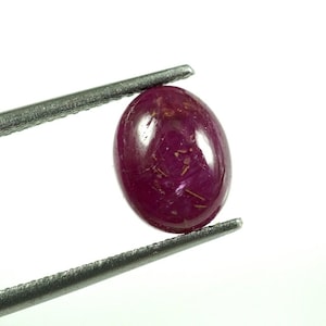 Op de afbeelding: Een ovaalvormige, diep magentarode edelsteen met een glad, gepolijst cabochon-oppervlak. De steen vertoont zichtbare interne insluitingen en een natuurlijke textuur, vastgehouden tussen de twee metalen uiteinden van een pincet.