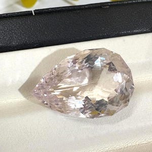 May include: A pear-shaped, faceted gemstone with a pale pink hue. The stone features a brilliant cut with numerous triangular and kite-shaped facets that reflect light, creating a sparkling appearance. The gem is displayed in a jewellery box.