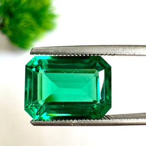Pode incluir: Uma gema de cor verde esmeralda vibrante, lapidada em formato retangular com facetas escalonadas. A pedra é mantida firmemente por uma pinça de metal prateado, exibindo sua superfície transparente e sua cor verde intensa e saturada.