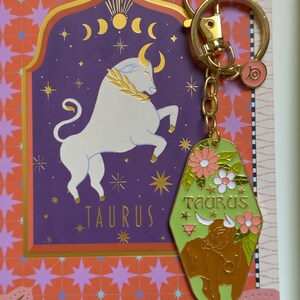 May include: A gold-toned keychain with a green and gold Taurus-themed charm. The charm features a brown bull illustration, flowers, and the word "TAURUS". The background includes a purple Taurus sign and a gold-colored ring.