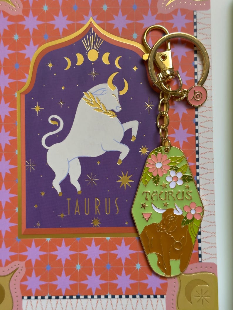 May include: A gold-toned keychain with a green and gold Taurus-themed charm. The charm features a brown bull illustration, flowers, and the word "TAURUS". The background includes a purple Taurus sign and a gold-colored ring.