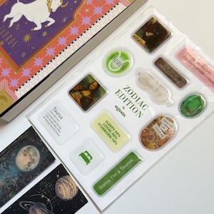 May include: A Zodiac Edition sticker sheet and box set featuring Taurus imagery. The box has a purple and orange design with a bull illustration. The sticker sheet includes various designs, such as the Mona Lisa, a sleep mask, and text that reads "Sorry, I'm a Taurus."