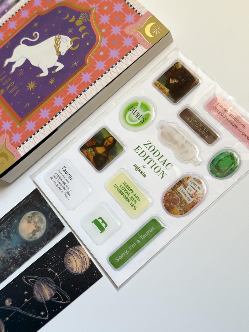 May include: A Zodiac Edition sticker sheet and box set featuring Taurus imagery. The box has a purple and orange design with a bull illustration. The sticker sheet includes various designs, such as the Mona Lisa, a sleep mask, and text that reads "Sorry, I'm a Taurus."