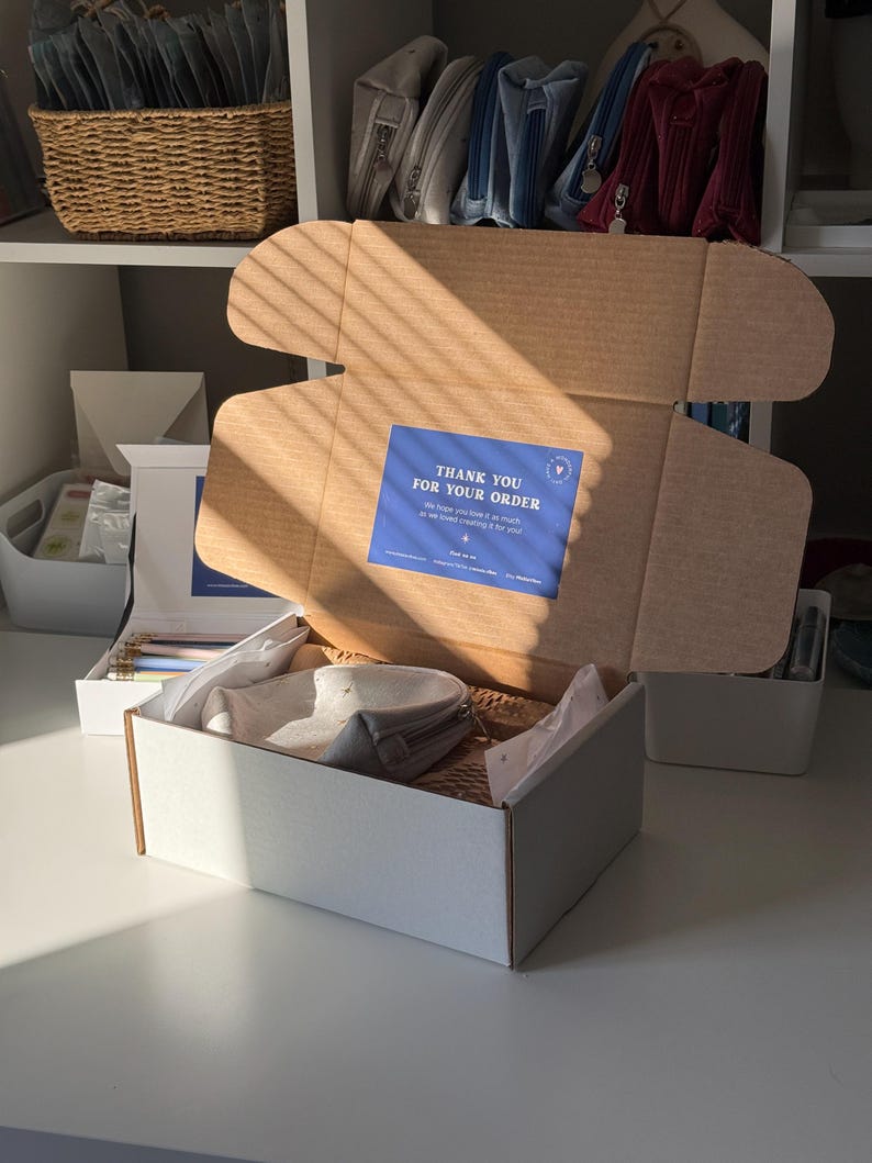 May include: An open cardboard box with a blue sticker that reads "THANK YOU FOR YOUR ORDER". Inside the box are gray pouches and tissue paper. The box is on a white surface, with a shelf and other items in the background.