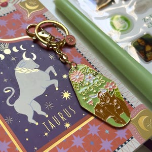 May include: A gold-colored keychain with a green and floral Taurus design. The keychain is attached to a purple and gold patterned notebook. A green candle and other small items are also visible.