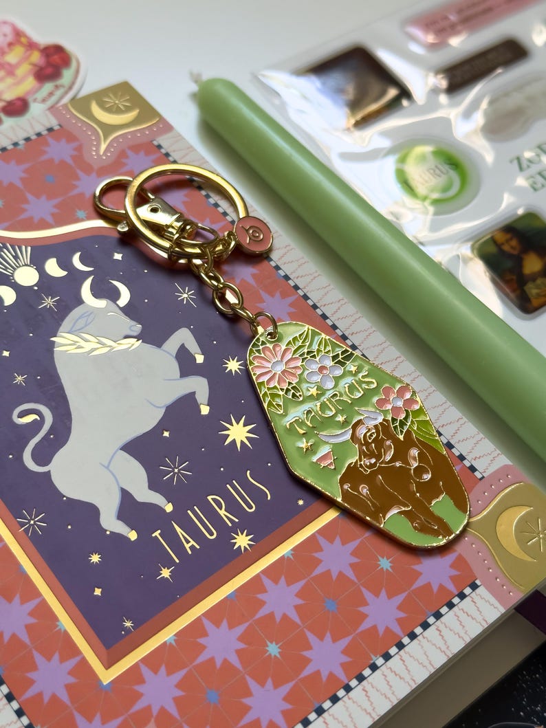 May include: A gold-colored keychain with a green and floral Taurus design. The keychain is attached to a purple and gold patterned notebook. A green candle and other small items are also visible.