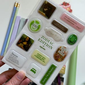 May include: A sheet of Zodiac Edition stickers with various designs and text. The stickers feature images, phrases like "I'm not stubborn, I'm passionate - okay?", and the Taurus symbol. The sheet is held by a hand.