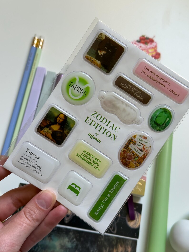 May include: A sheet of Zodiac Edition stickers with various designs and text. The stickers feature images, phrases like "I'm not stubborn, I'm passionate - okay?", and the Taurus symbol. The sheet is held by a hand.