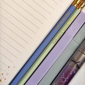 May include: An assortment of stationery items including a lined notepad, pencils, and highlighters. The pencils are blue and green with gold text that reads "FREE TO BE YOU" and "THOUGHTS SHAPE VISION." A clear container with the word "Astrology" and a Taurus graphic is also visible.