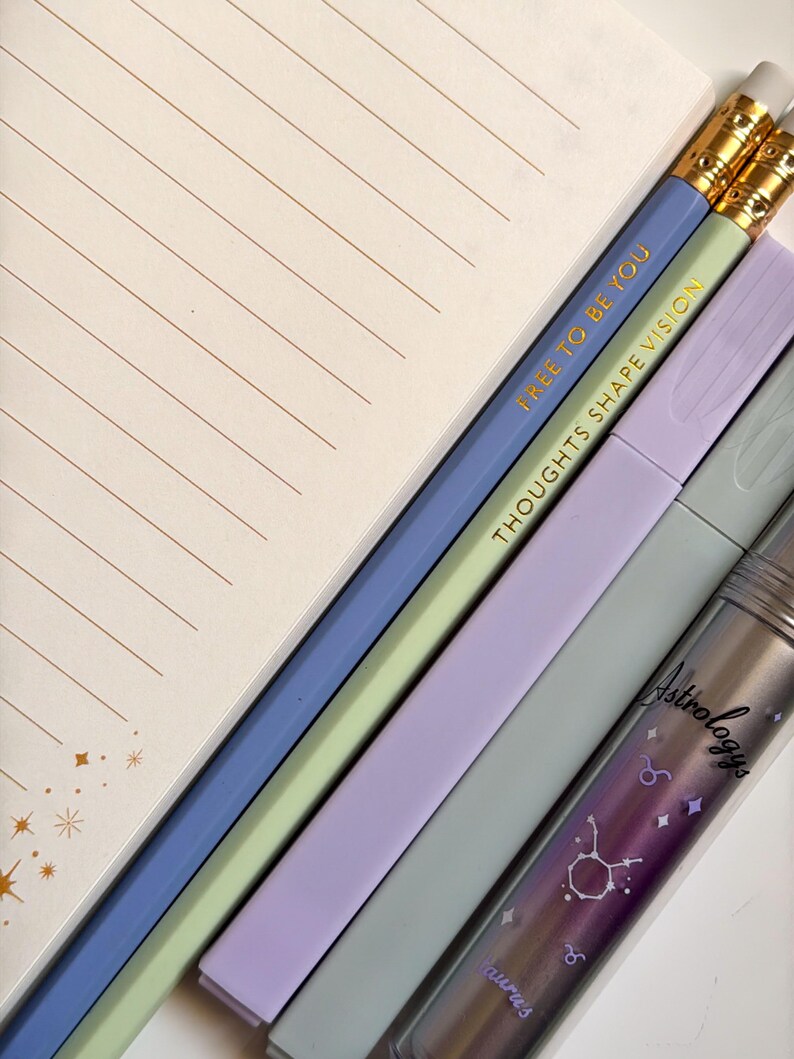 May include: An assortment of stationery items including a lined notepad, pencils, and highlighters. The pencils are blue and green with gold text that reads "FREE TO BE YOU" and "THOUGHTS SHAPE VISION." A clear container with the word "Astrology" and a Taurus graphic is also visible.