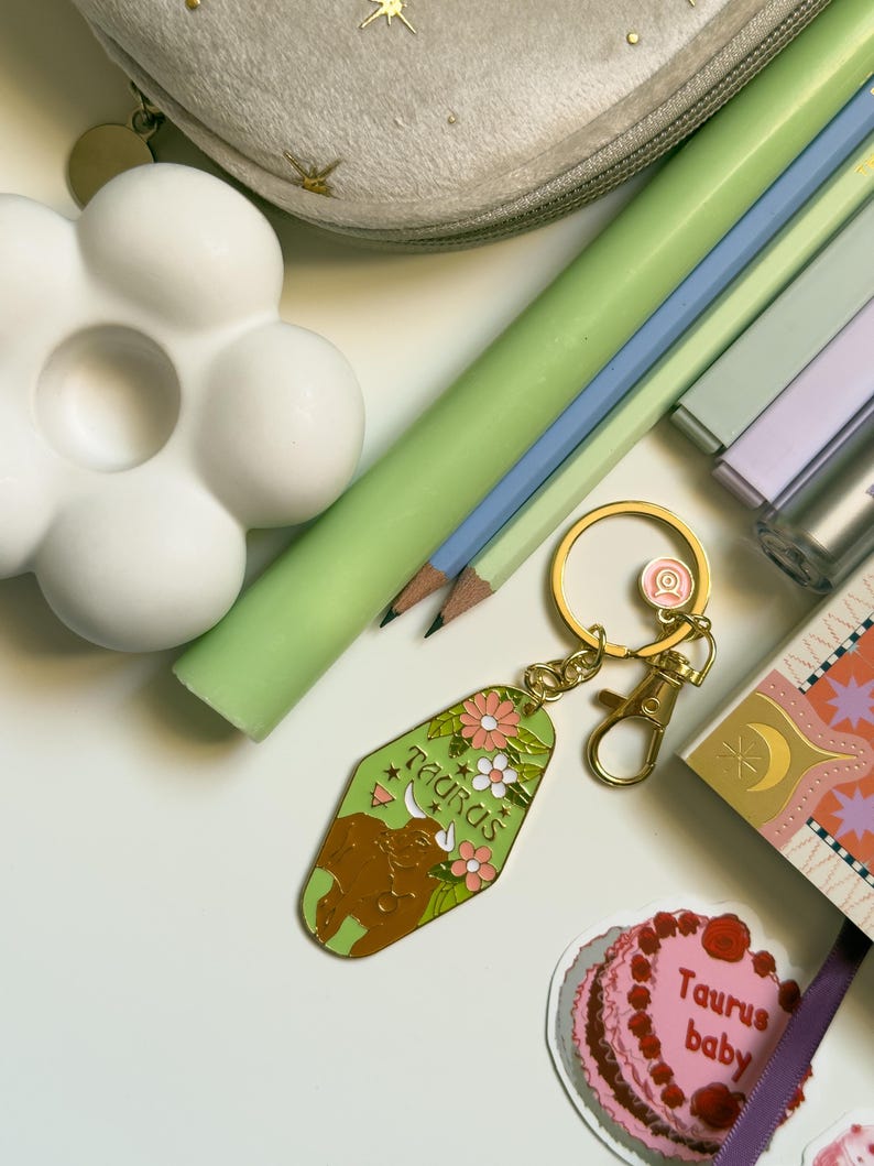 May include: A collection of stationery items including pencils, a flower-shaped object, and a Taurus keychain. The keychain is green with a brown bull illustration and the word "Taurus". A small cake sticker with the words "Taurus baby" is also visible.