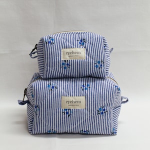 Peut inclure: Deux trousses de maquillage à rayures bleues et blanches superposées. Les sacs ont un motif matelassé avec de petits accents floraux bleus. Chaque sac a une fermeture éclair et une étiquette "ryehem".