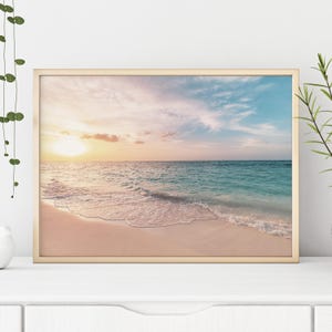 Peut inclure: Une impression encadrée d'une scène de plage au coucher du soleil. L'image présente un cadre doré, une plage de sable et une eau turquoise sous un ciel aux teintes roses, oranges et bleues. Le soleil se couche à l'horizon.