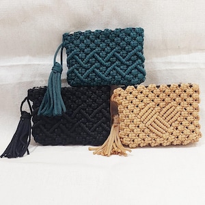 Op de afbeelding: Drie macramé clutch portemonnees in teal, zwart en tan. Elke rechthoekige portemonnee heeft een geometrisch patroon en een bijpassende kwast. De portemonnees worden gepresenteerd op een witte achtergrond, waarbij de handgemaakte details en kleurvariaties worden getoond.