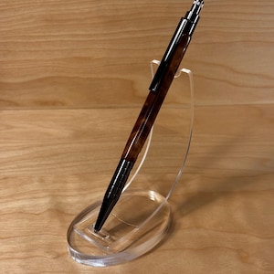 Puede incluir: Un bolígrafo marrón y negro con un patrón de vetas de madera se exhibe en un soporte de acrílico transparente. El bolígrafo tiene una punta y detalles metálicos brillantes. El bolígrafo está en ángulo, mostrando su diseño.