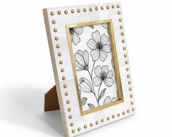 May include: A rectangular white photo frame with a gold inner frame and gold-colored studs around the border. The frame holds a black and white floral print. The frame is propped up with a gold-colored stand.