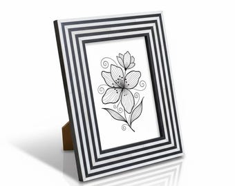 Puede incluir: Un marco de fotos a rayas blancas y negras con un dibujo lineal floral. El marco está en ángulo y sostenido por un caballete de madera. La obra de arte presenta una flor detallada con pétalos y hojas arremolinadas.