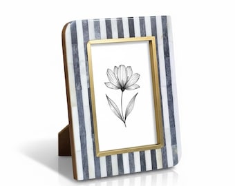 Puede incluir: Un marco de fotos rectangular con un patrón de rayas en tonos grises y blancos. El marco tiene un borde interior dorado y un soporte de madera. Contiene una ilustración floral en blanco y negro.