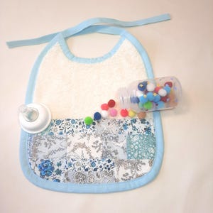 Può includere: Un bavaglino per bambini con bordi e lacci azzurri. Il bavaglino ha una parte superiore in spugna bianca e una sezione inferiore floreale blu fantasia. Una bottiglia di plastica trasparente piena di pompon colorati è accanto al bavaglino.