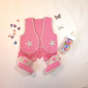 May include: A pink crochet baby vest with fringe and star accents, paired with matching boots. The vest has a light gray trim and two star appliqués. The boots have a light gray top and a small flower detail. A charming baby outfit.