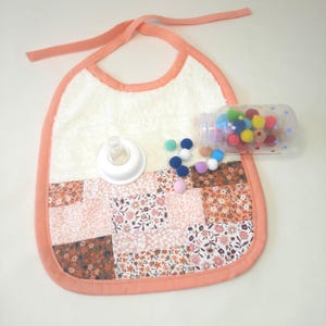Puede incluir: Babero para bebé con diseño patchwork floral en tonos rosas y marrones, ribeteado con tela melocotón. Cuenta con una sección superior de rizo blanco, una tetina y un biberón transparente lleno de pompones de colores.