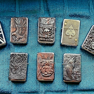 Op de afbeelding: Een verzameling van zeven metalen aanstekers met verschillende reliëfontwerpen. De ontwerpen omvatten een adelaar, een stammasker, een hert, een bulldog, een vis, een vrouwengezicht en een Union Jack-vlag. De aanstekers zijn in zilver, brons en goud.