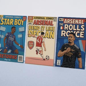 May include: Three Arsenal comic book style prints. The prints feature illustrations of soccer players with text that reads "STAR BOY RETURNS", "BEND IT LIKE DECAN", and "THE ROLLS ROYCE". The prints have a vintage comic book aesthetic.