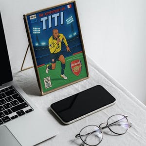 May include: A framed comic book cover featuring a cartoon illustration of a soccer player in a blue and yellow jersey with the text "THE ADVENTURES OF TITI" and "Arsenal" on the cover.