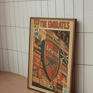 Puede incluir: Póster enmarcado con el logotipo del club de fútbol Arsenal y el texto "The Emirates". El póster tiene un estilo de cómic vintage con tonos rojos, amarillos y marrones. El texto "Arsenal Maldives" y "Germany" está incluido.