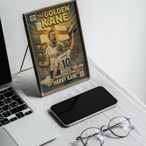 Puede incluir: Obra de arte enmarcada estilo c&oacute;mic con el texto "The Golden Kane" y "There's Only One Harry Kane". La obra representa a un jugador de f&uacute;tbol sosteniendo un trofeo. Tambi&eacute;n se ven un port&aacute;til, un tel&eacute;fono y gafas.
